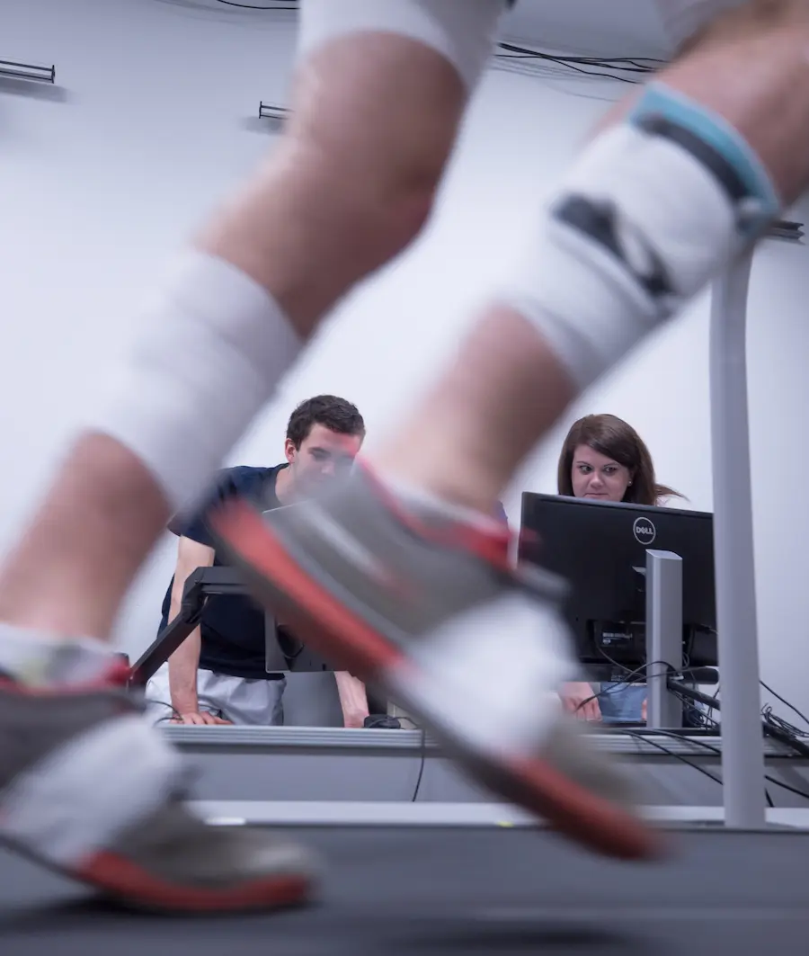 Close up of legs running on a treadmill while people observe metrics on a computer